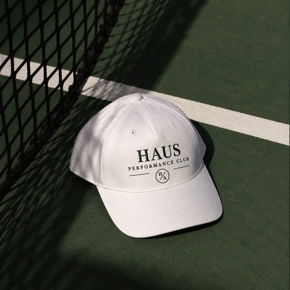 White HAUS Performance Club cap with green embroidery resting on a green court beside a net
